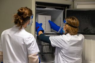 White box of Pfizer-BioNTech vaccines being placed in a freezer by two gloved scientists in lab coats