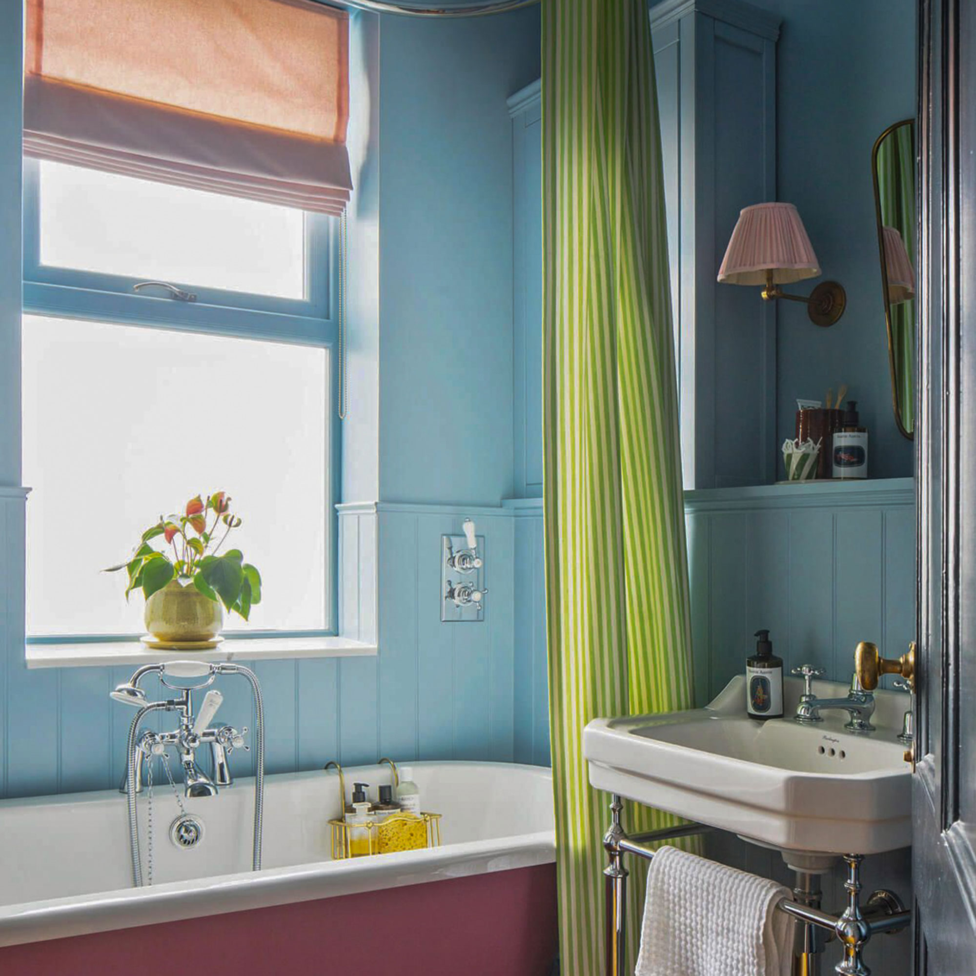 Blue panelled bathroom with roll top bath, green stripy shower curtain and traditional pedestal style sink