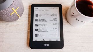 The Kobo Clara 2E laying flat on a table next to a speaker and coffee mug, showing the collection of books loaded onto the device.