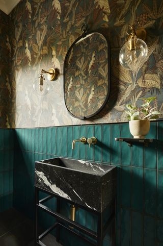 Image of a dark, art deco-inspired bathroom. There is dark teal tile on half of the wall and dark teal, gold, and brown tropical wallpaper on the other half. The sink is a black marble material and there is an oval black mirror hanging above it.