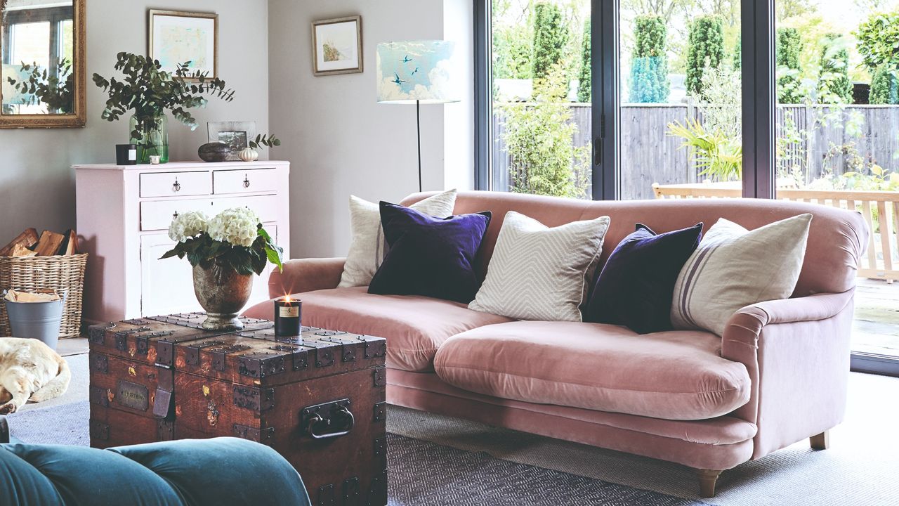 A living room with a pink velvet sofa adorned with two cream linen cushions with a stripe, two purple velvet cushions and a cream cushion in the middle