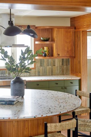 Mid-century modern kitchen with warm wood cabinetry and a matching wooden island. There are colorful terrazzo worktops on both the counter and the island. The cabinets are sage green and there is sage green tile on the backsplash.