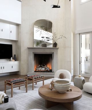 Large mirror on modern concrete fireplace in living room with gray rug, round wooden coffee table and decorative stools