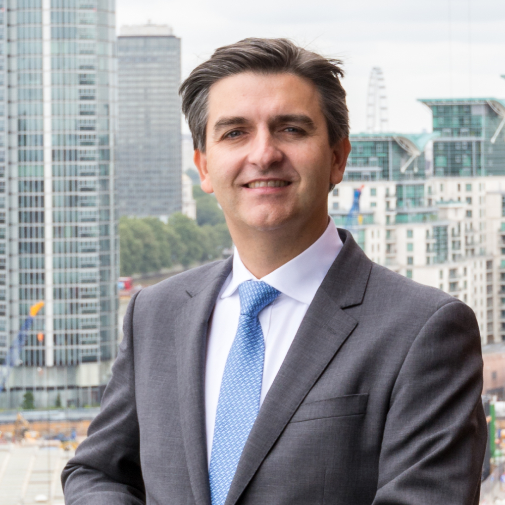 man with short dark hair in grey suit, white shirt and blue tie, stood with London skyline in background 