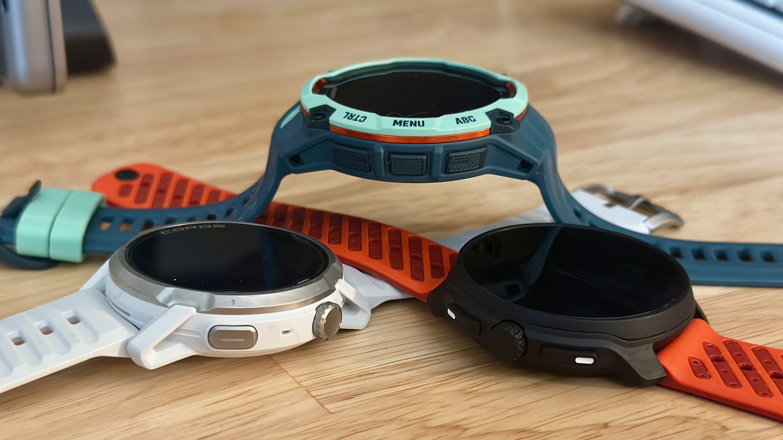 A Garmin Instinct 3 AMOLED sitting propped up on its strap above the COROS APEX 4 (left) and Suunto Race 2 (right) at diagonal angles, placed to emphasize their rotating crowns compared to Garmin's buttons.