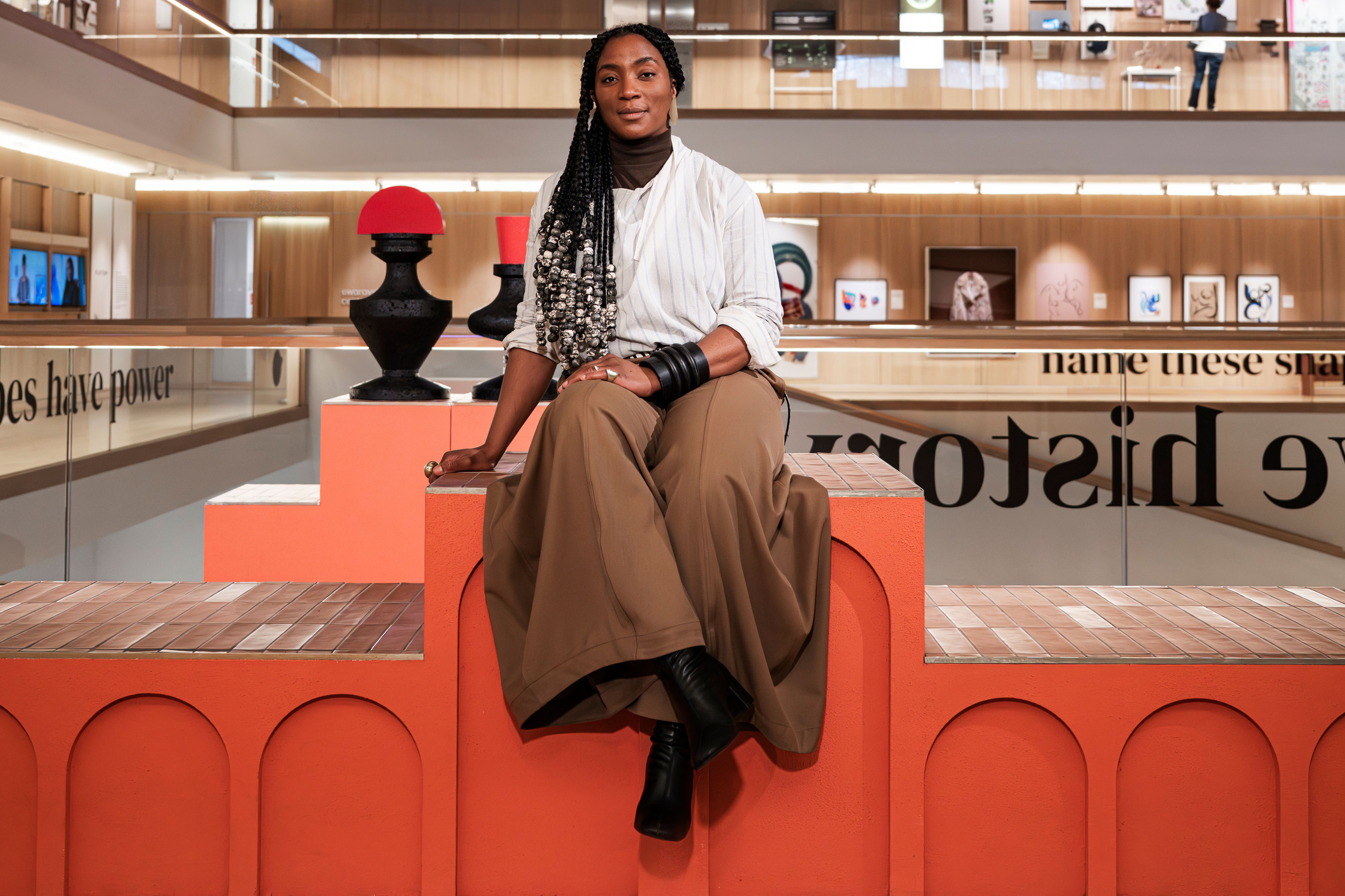 A young woman dressed in a white stripy shirt worn over a brown turtleneck, loose brown trousers, black leather jewelry, and boots sits on an orange-painted wooden podium in a brightly lit gallery.
