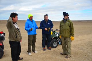Four people stand in the barren arctic tundra alongside ATVs.