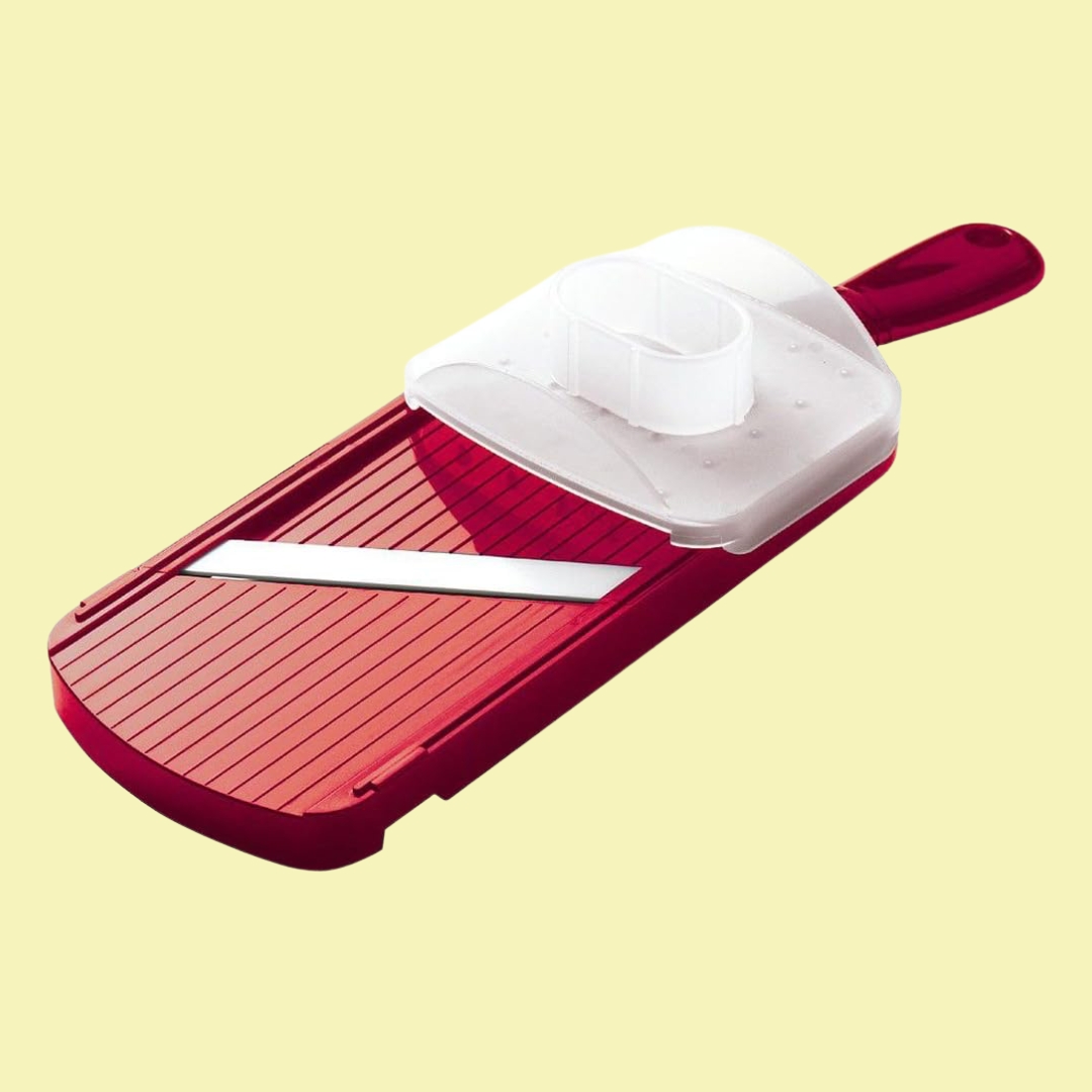 A red plastic mandolin with white ceramic blade sits against a pale yellow background in diagonal position.