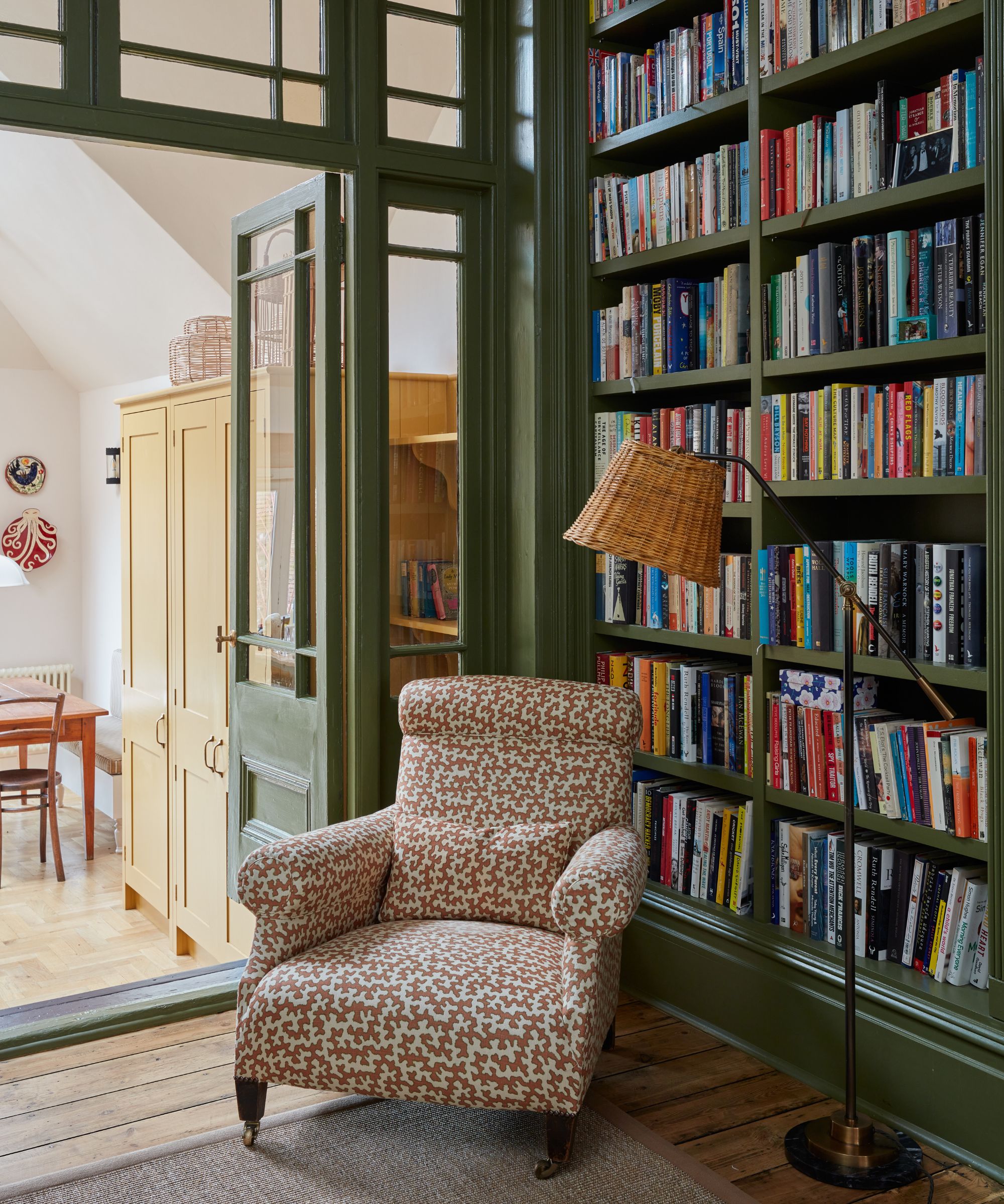 Home library with built-in green bookshelves filled with books, a rattan floor lamp, an upholstered patterned pink armchair, wooden flooring, looking into a bright kitchen with yellow cabinetry.