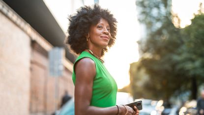 best serums for mature skin - black woman with Afro hair smiling softly, wearing sleeveless green slim-fitting dress and red lipstick and gold jewellery, holding her phone 