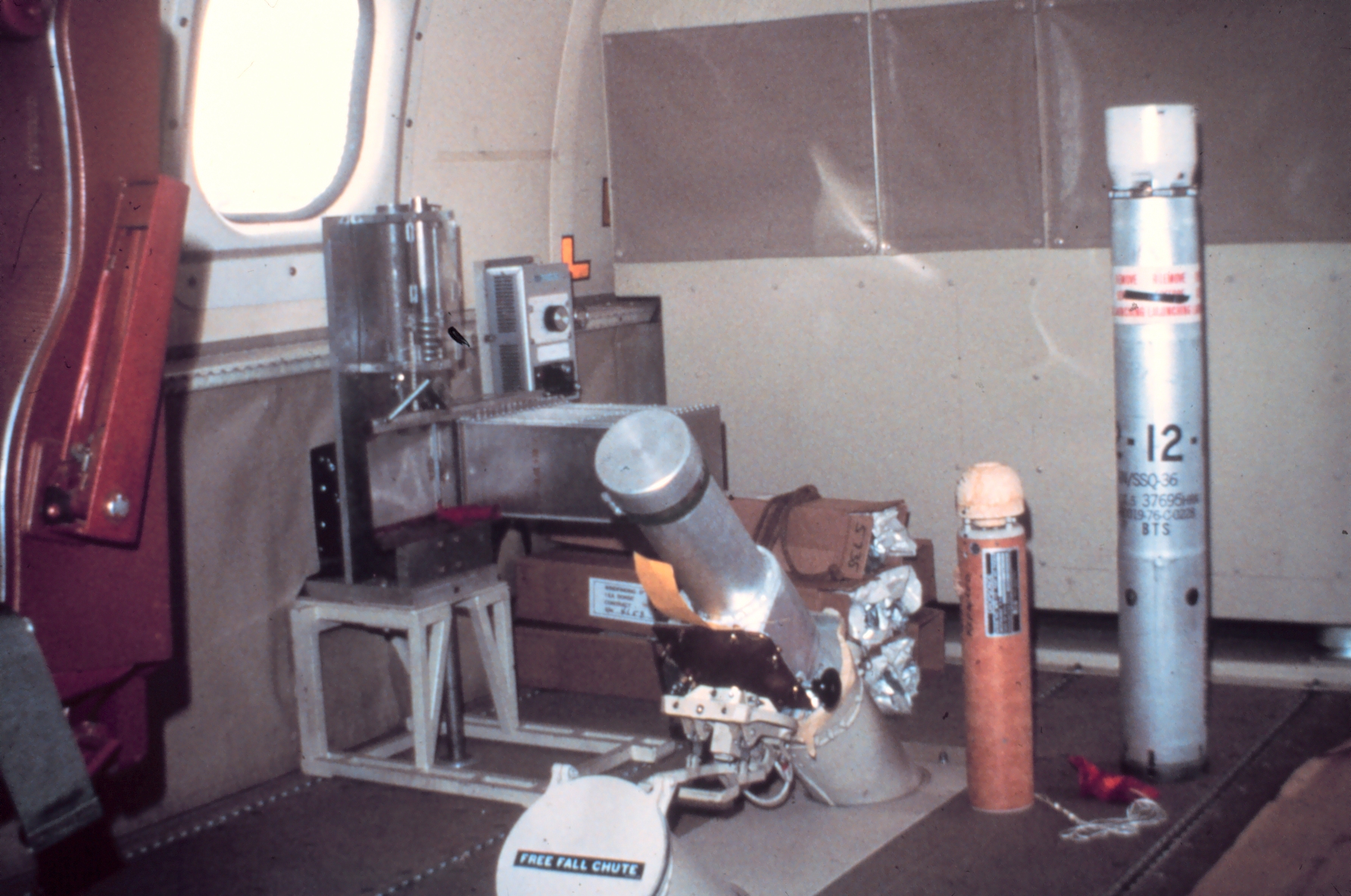 inside an aircraft cabin, a small metal cylinder waits atop a chute that leads through the floor