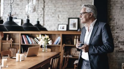 An older businessman holding a coffee cup stands in his office and gazes calmly into the distance.