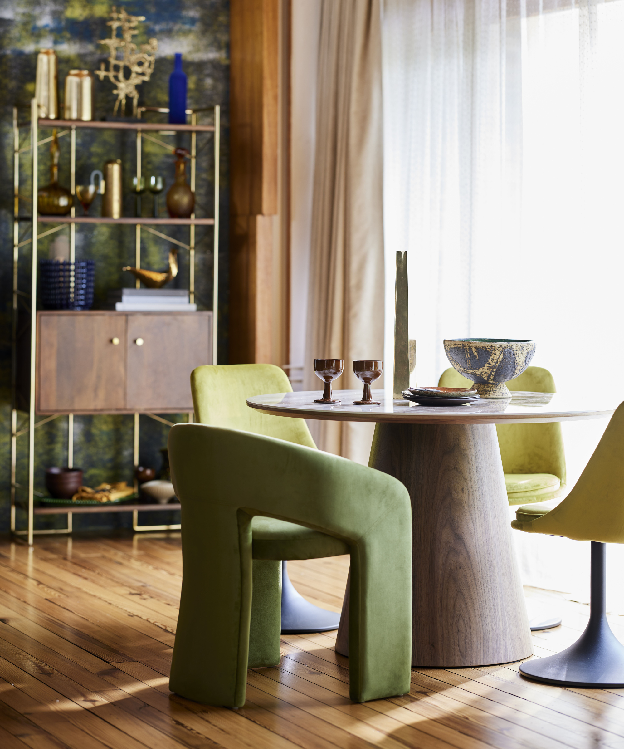 round modern wooden dining table with velvet modern chairs, large windows, curtains and shelving unit against wallpapered wall