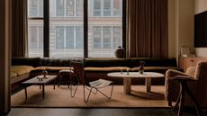 living room with slow-slung sofa, two coffee tables, an ottoman, slung chair, boucle upholstered armchair, and deep orange colored floor rug, with heavy curtains covering large windows