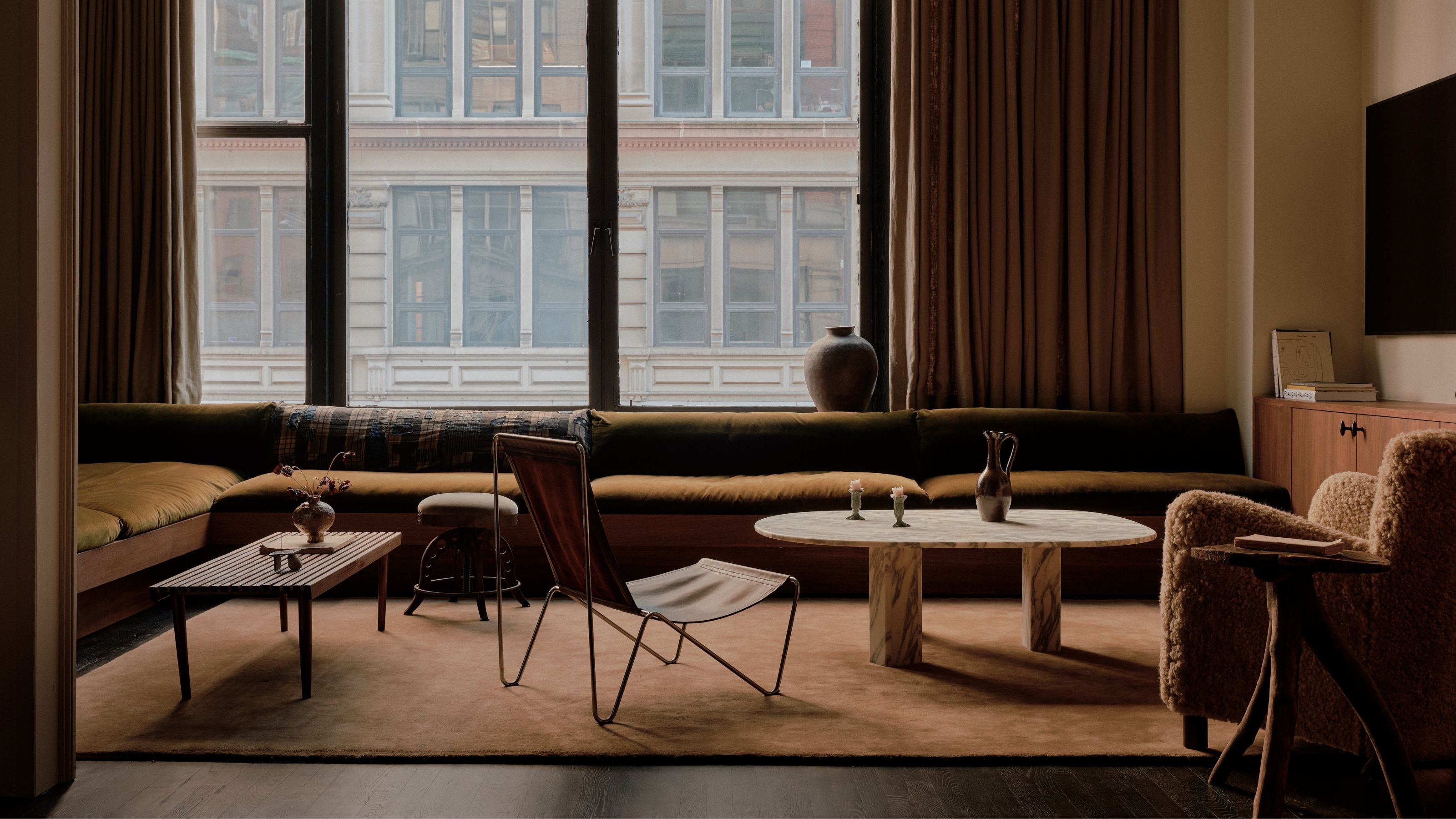 living room with slow-slung sofa, two coffee tables, an ottoman, slung chair, boucle upholstered armchair, and deep orange colored floor rug, with heavy curtains covering large windows
