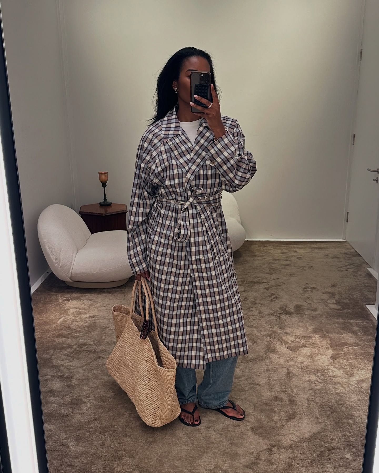 A woman wearing a plaid trench coat, a large woven bag, jeans, and black flip flops.