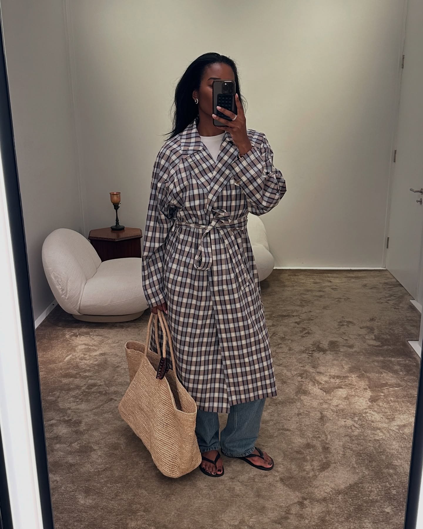 A woman wearing a plaid trench coat, a large woven bag, jeans, and black flip flops.