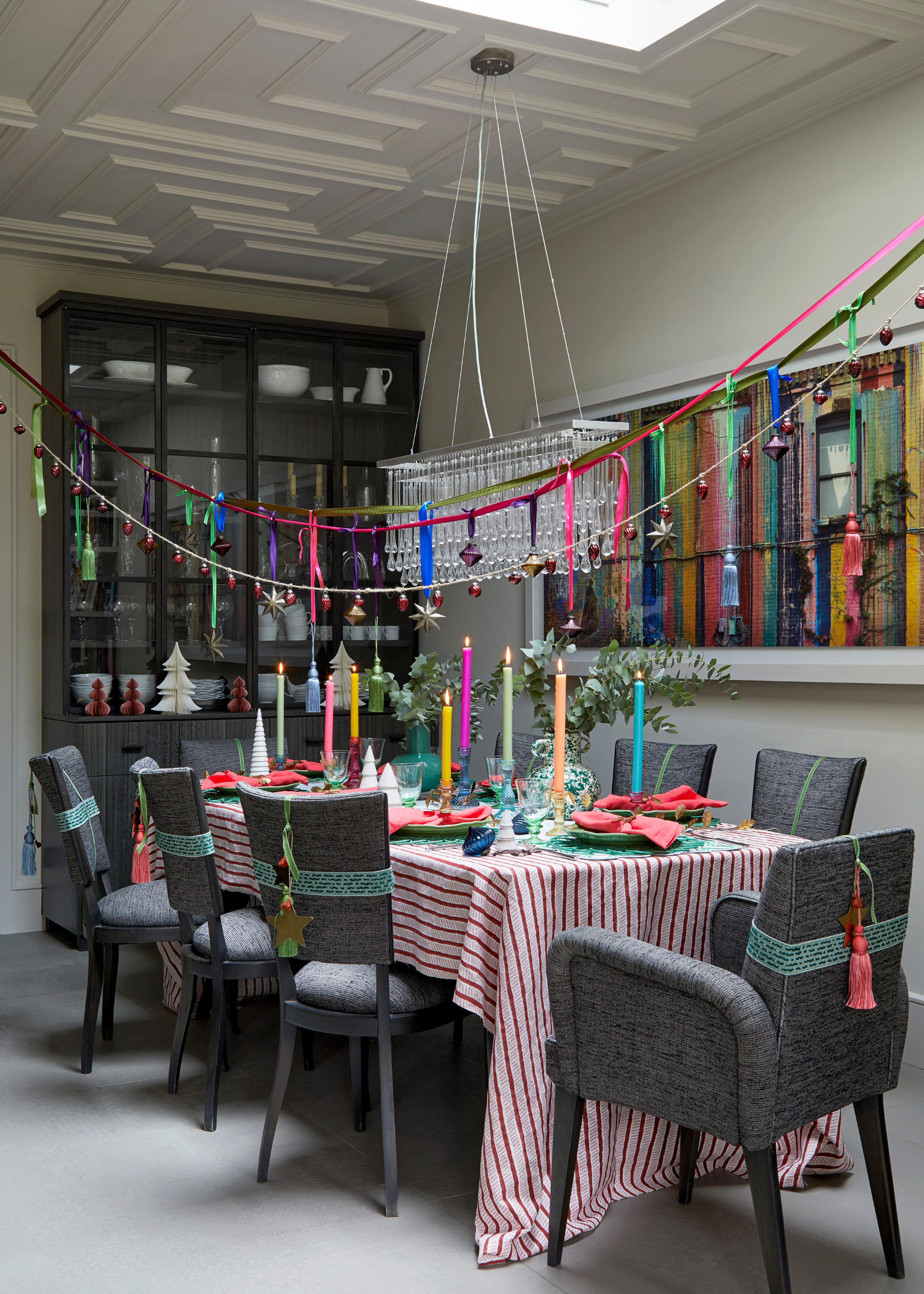 A dining table decorated for Christmas with hanging string lights and ribbons, a striped tablecloth that drapes onto the floor, colorful tassles on the backs of the dining chairs and an array of multicolored taper candles on the table