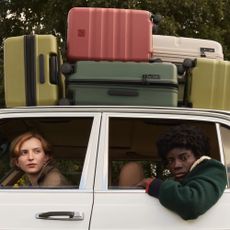An image of the side of the car with a woman looking out of the front window and another woman hanging her arm out of the back window with several Antler suitcases on top of the car