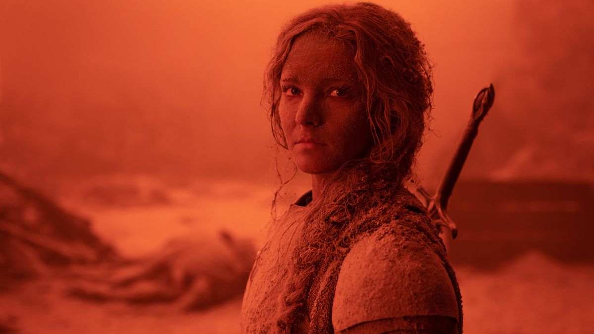 An armored Galadriel is covered in dirt and ash in a red-lit location as she stares into the camera in The Rings of Power