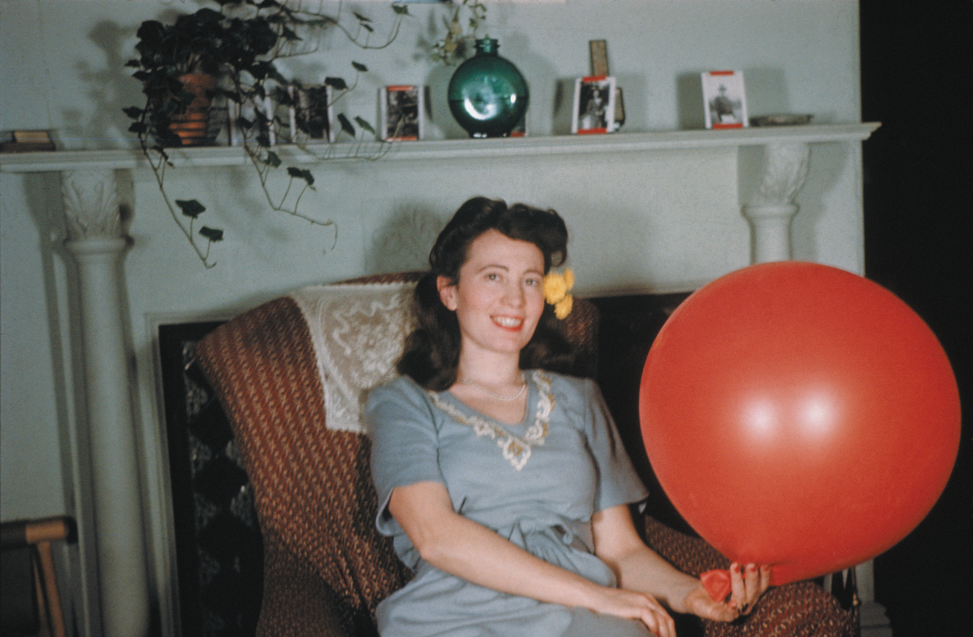 A color photograph of a smiling woman sitting in an armchair next to a large red balloon, with a yellow flower in her hair.