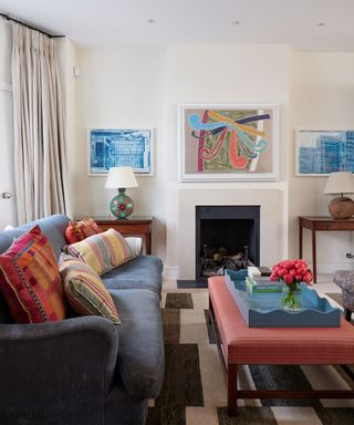 Bright living room with cream painted walls, fireplace, artwork mounted either side on wall, two wooden console tables, blue sofa, red ottoman, blue tray with flowers, artwork above fireplace