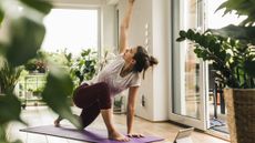 A woman performs a modified version of the World's Greatest Stretch in a light, airy living room filled with leafy plants