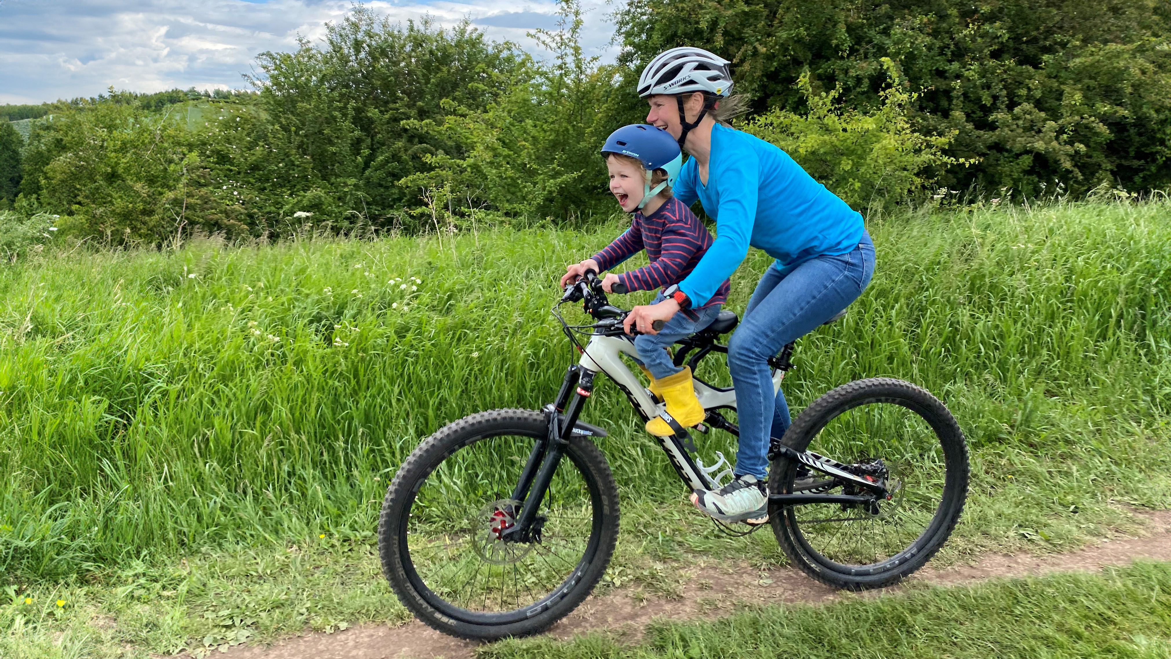 Mum riding a mountain bike with her toddler sat on a kids ride shogun pro seat, smiling and laughing as they wizz along. 