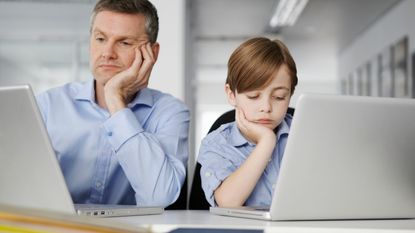 A father and son in matching blue shirts have matching bored expressions as they look at laptops.