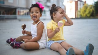 two young girls sit on the ground outside an apartment building eating ice cream. One of the girls is holding her head as if experiencing brain freeze.