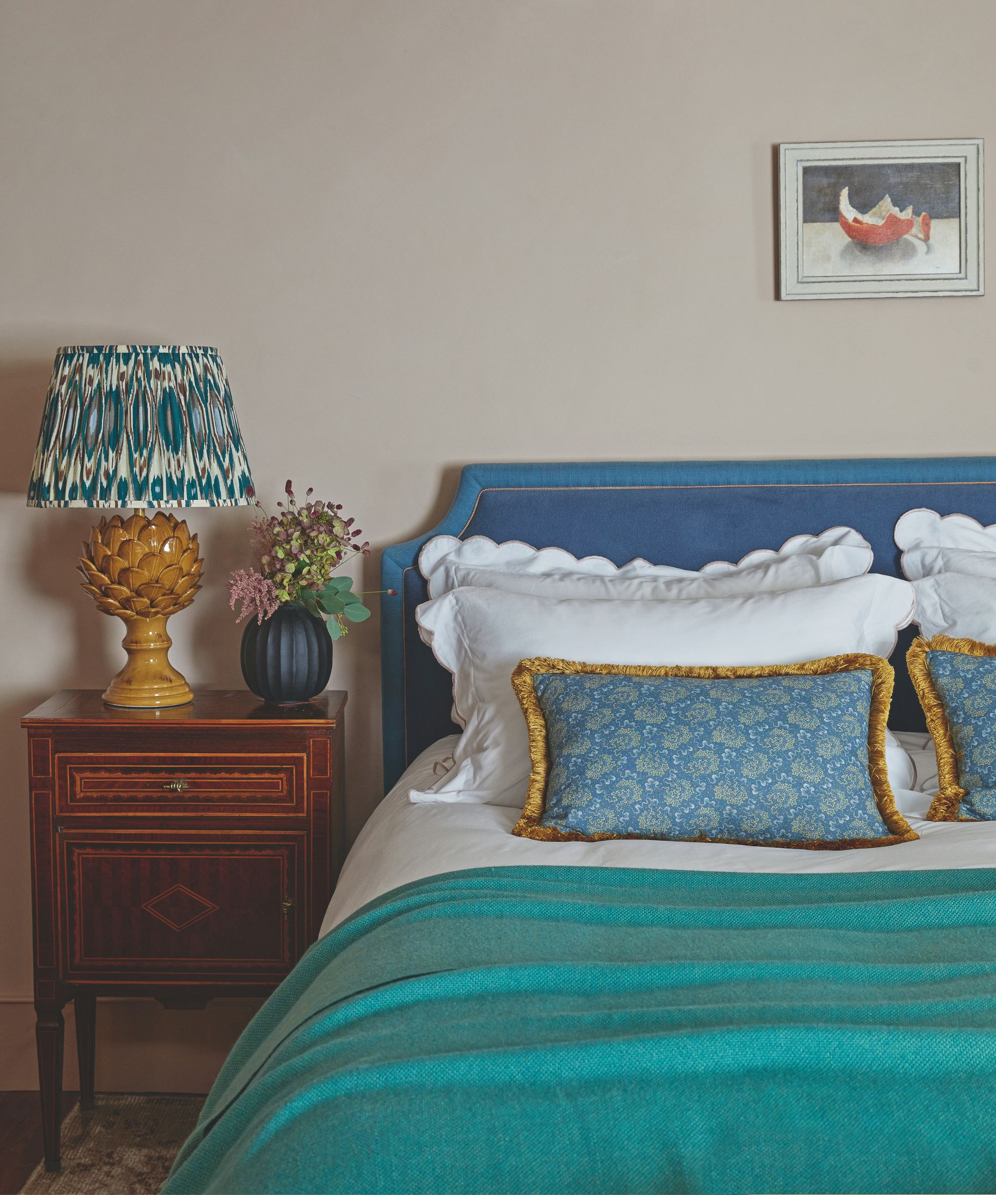 A bedroom with a bright blue bed with blue bedding, a wooden bedside table, statement yellow ceramic lamp with patterned shade, small blue vase with flowers, neutral walls and a small still life artwork above the bed