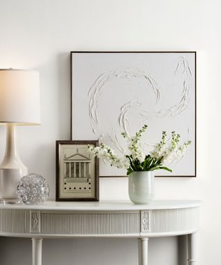 white wall with white console table, white textured artwork in thin black frame, table lamp, framed photo and white and green flowers in a vase