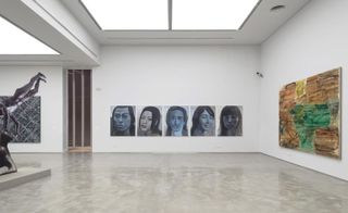 Art exhibition space with white walls. On the far wall, a long piece of artwork displays 5 portrait images in different shades of blue and grey. blue.
