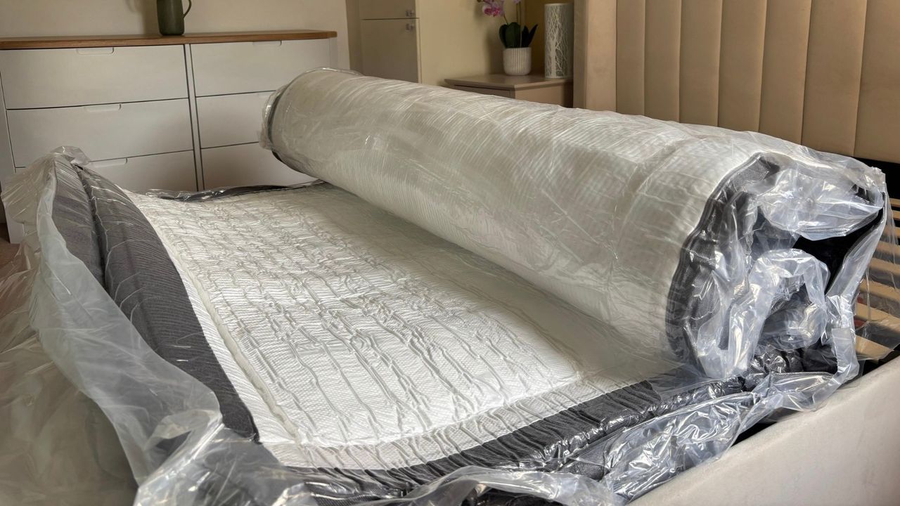 The image shows a dark grey and white mattress on a grey bed frame expanding inside its plastic wrapping