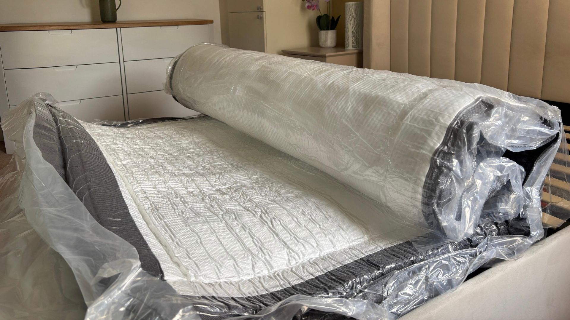 The image shows a dark grey and white mattress on a grey bed frame expanding inside its plastic wrapping