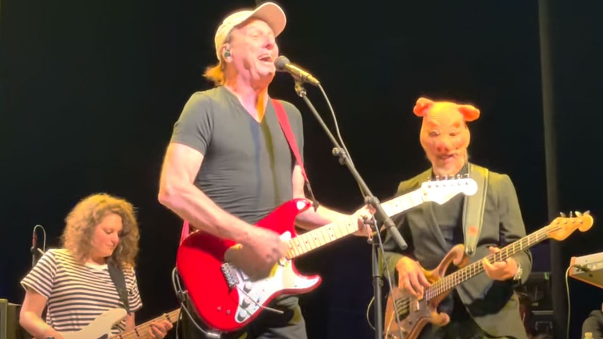 Adrian Belew playing guitar onstage with bassist Julie Slick and Les Claypool in a pig&#039;s mask