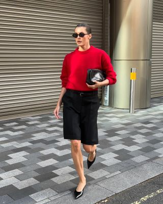 A influenciadora de moda britânica Valeria @sobalera em uma calçada de Londres usando óculos escuros ovais pretos, um suéter vermelho de gola redonda, bolsa clutch preta, bermuda preta com cinto e saltos gatinho pretos