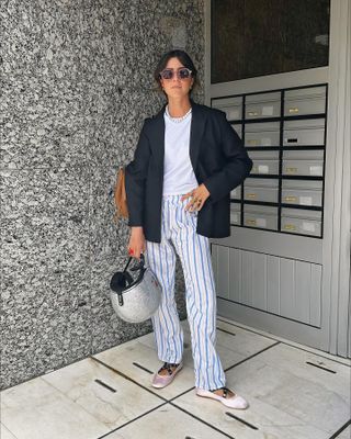 Spanish influencer Vicky Montanari poses outside a building holding a scooter helmet while wearing a beaded necklace, black blazer jacket, white T-shirt, blue striped pajama pants, and pink Miu Miu ballerina flats.