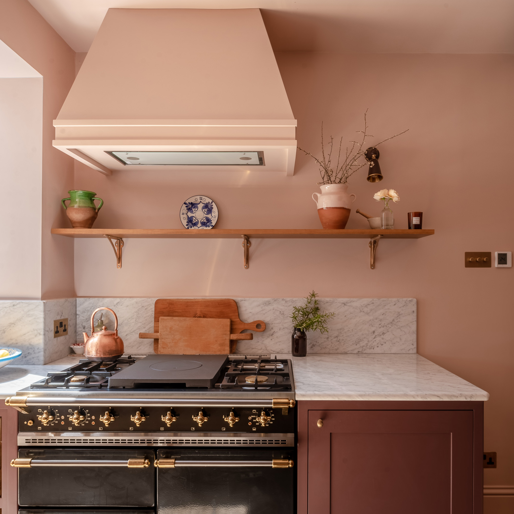 close up of black range cooker in kitchen with pink walls and pink painted extractor fan and purple cabinetry with marble worktop and splashback as well as an open shelf herringbone house