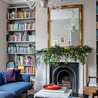 Sitting room with a Christmas mantel over the fire place below the mirror with a coffe table, blue sofa and a book rack as décor
