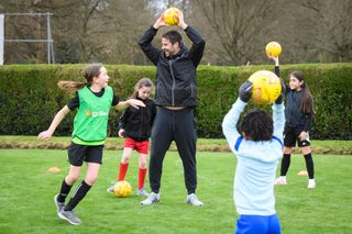 Former Liverpool and Tottenham midfielder Jamie Redknapp holds a ball above his head in a coaching session with young children