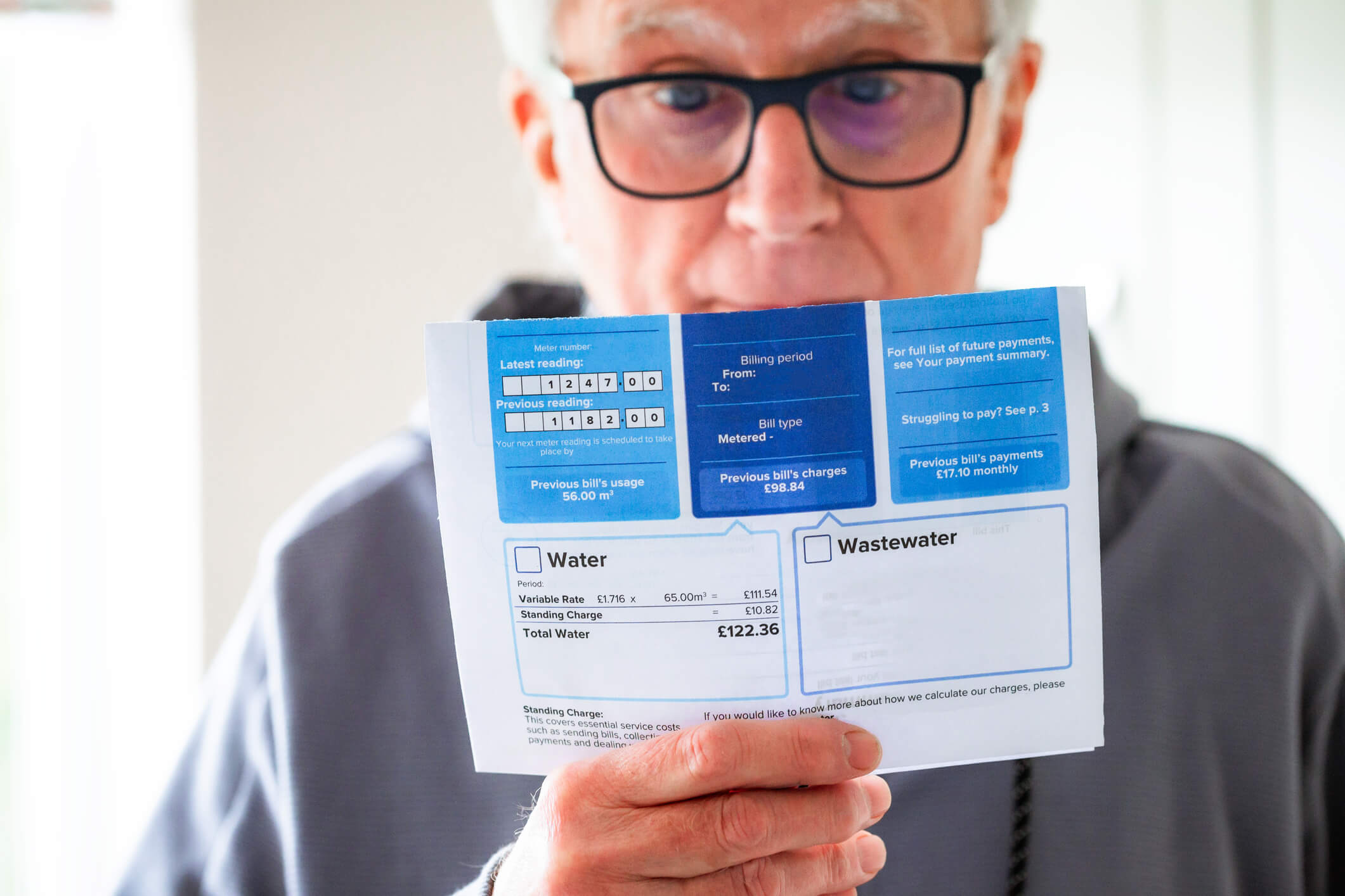 Portrait of a senior man in his 70s checking his water bill at home. He has a worried expression and touches his face with his hand while looking at the bill. Focus on the bill with the man&#039;s face defocused beyond.