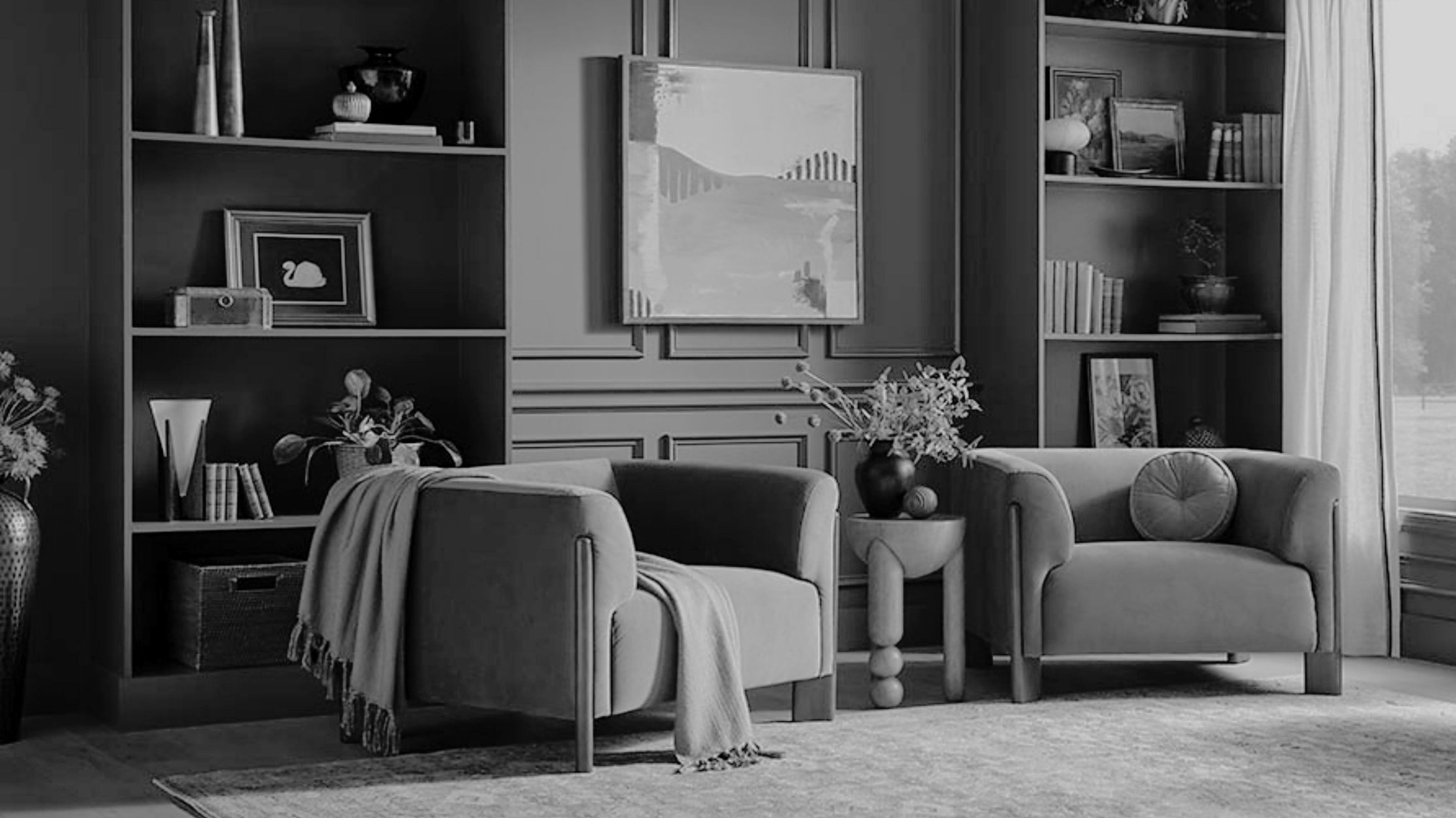 black-and-white image of a large living room with built-in bookshelves, two accent chairs, a rug, and windows with off-white curtains.