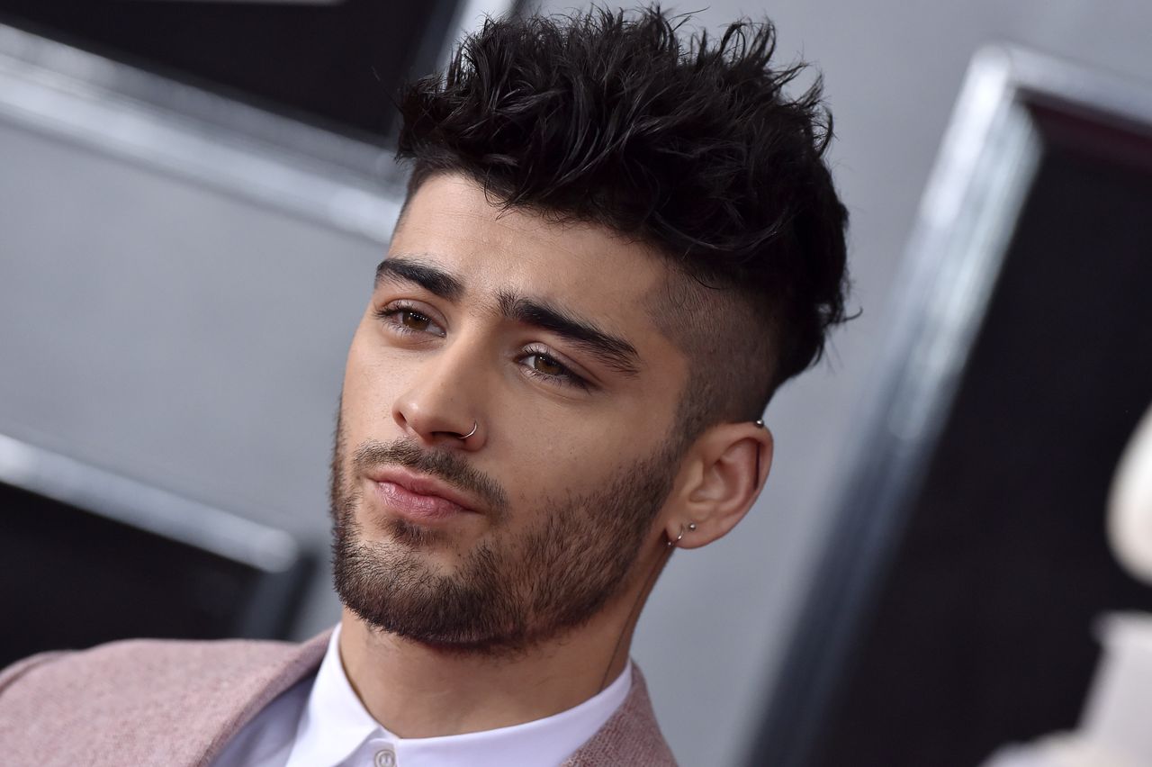 A headshot of Zayn Malik on a red carpet wearing a pink jacket and looking off-camera