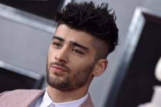 A headshot of Zayn Malik on a red carpet wearing a pink jacket and looking off-camera