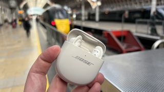 Bose QC Ultra Earbuds Gen 2 being held up in a train station with a train in the background