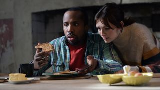 Mike (Kiell Smith-Bynoe) in a blue checked shirt and Alison (Charlotte Rtichie) in a grey jumped looked stunned at a slice of toast in the Button House kitchen in Ghosts. 