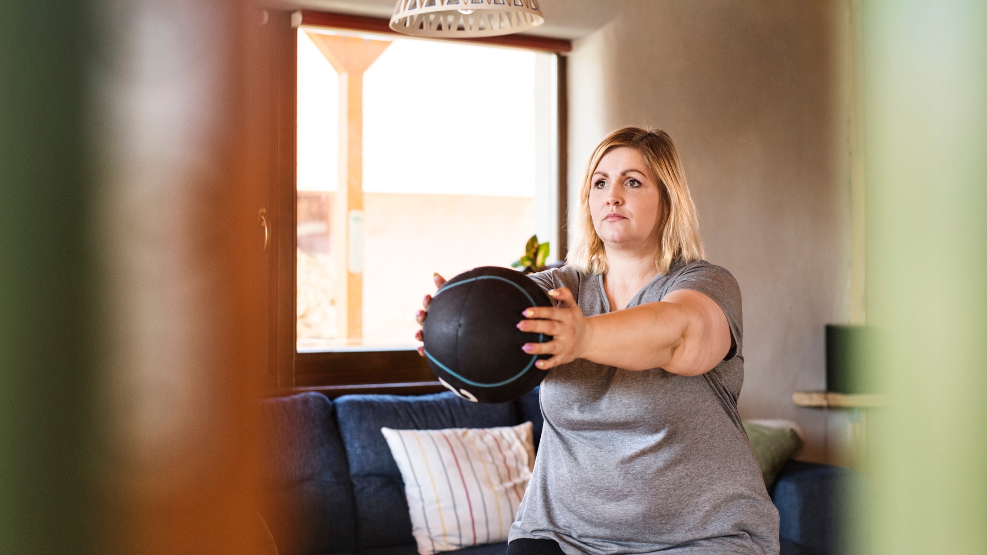 woman facing off camera standing holding a medicine ball arms outstretched in front. she's in a living room setting with a window behind her 