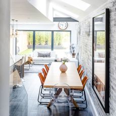 wooden brick wall room with a wooden dining table and orange chairs decorated with flowers in a vase next to a living room with a white sofa with patterned cushions and a white table topped with accessories 