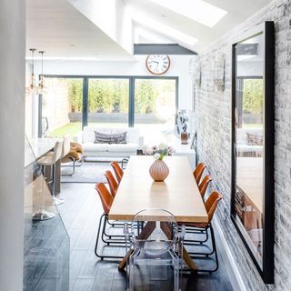 wooden brick wall room with a wooden dining table and orange chairs decorated with flowers in a vase, next to a living room with a white sofa with patterned cushions and a white table topped with accessories